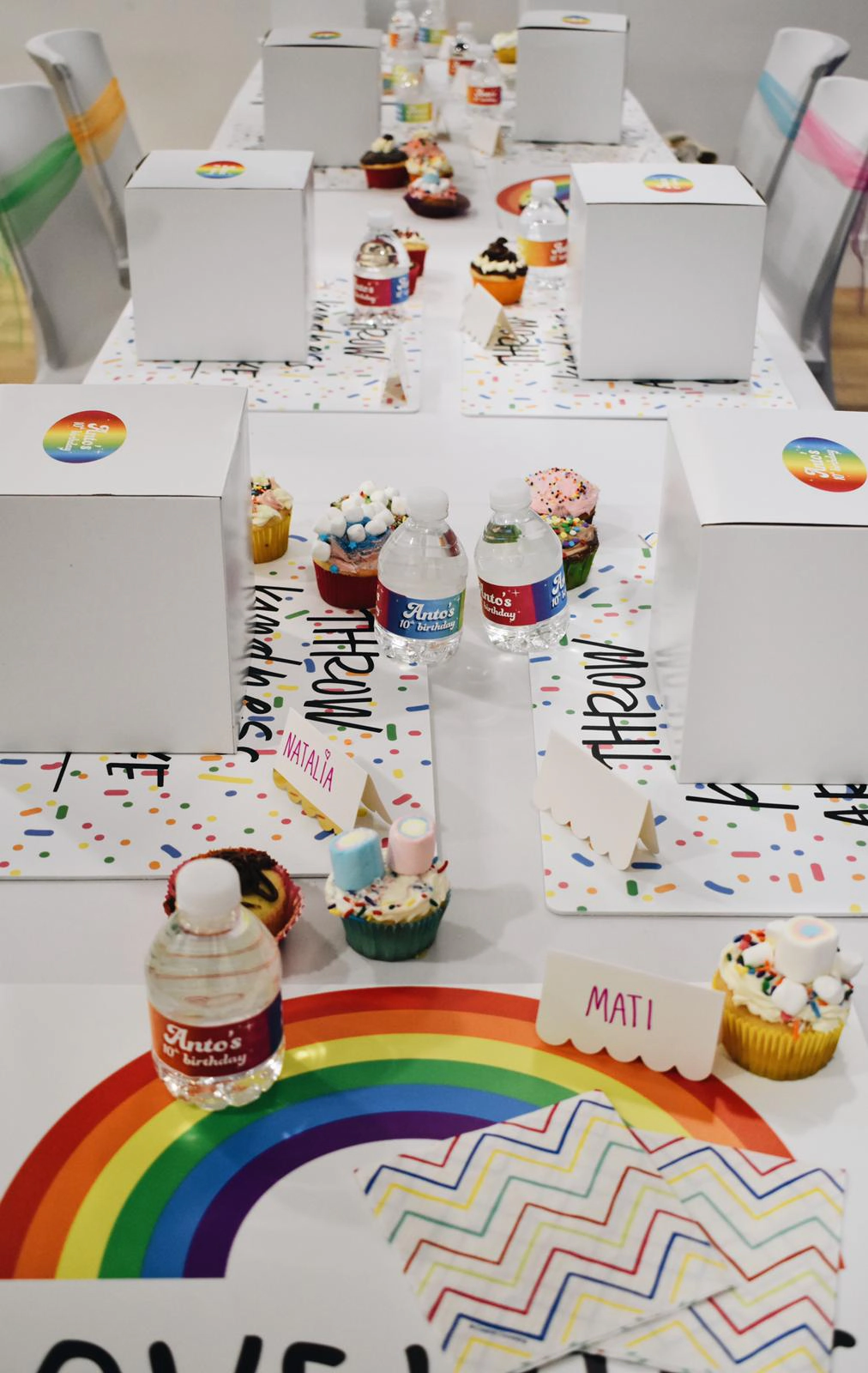 Long party table decorated with rainbow place mats, personalized water bottles, cupcakes with marshmallows, white favor boxes, and name cards