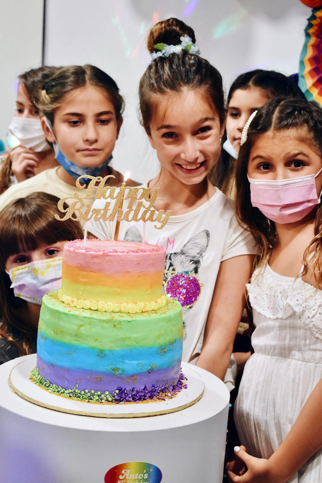 Smiling girl with a floral hair bun stands behind a two-tier rainbow birthday cake with a lit candle and gold “Happy Birthday” topper, surrounded by friends some wearing face masks at 10th birthday party
