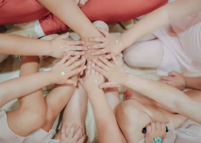 Overhead view of several children’s hands stacked together in a circle on the floor, each hand decorated with a small gold star sticker, showing friendship and teamwork in a dance class.