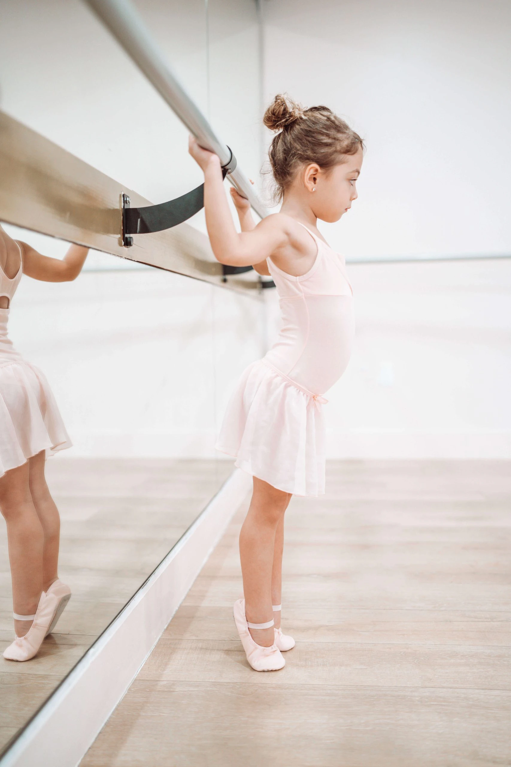 Young girl in a pale pink leotard, skirt, and ballet slippers rising onto her toes while holding the barre in front of a studio mirror