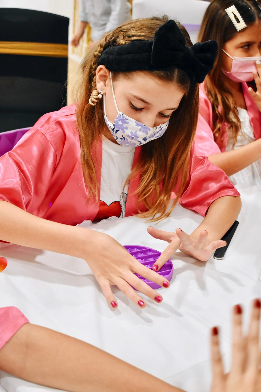 Young girl wearing a black bow headband, pink satin robe, and floral face mask presses a purple pop-it fidget toy on a white table during a kids’ spa party