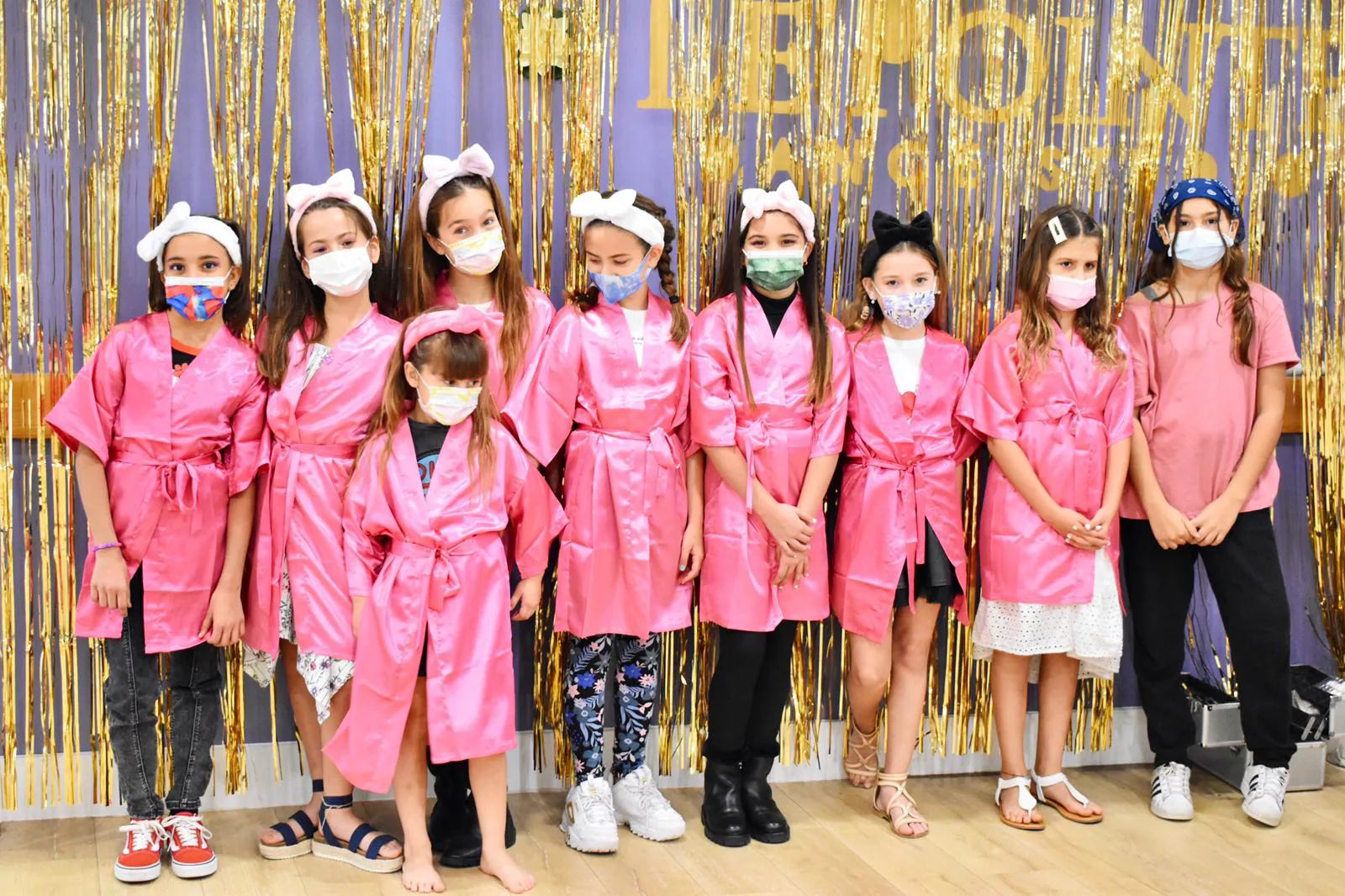 Group of nine girls wearing matching pink satin robes and various face masks pose together in front of a gold tinsel curtain
