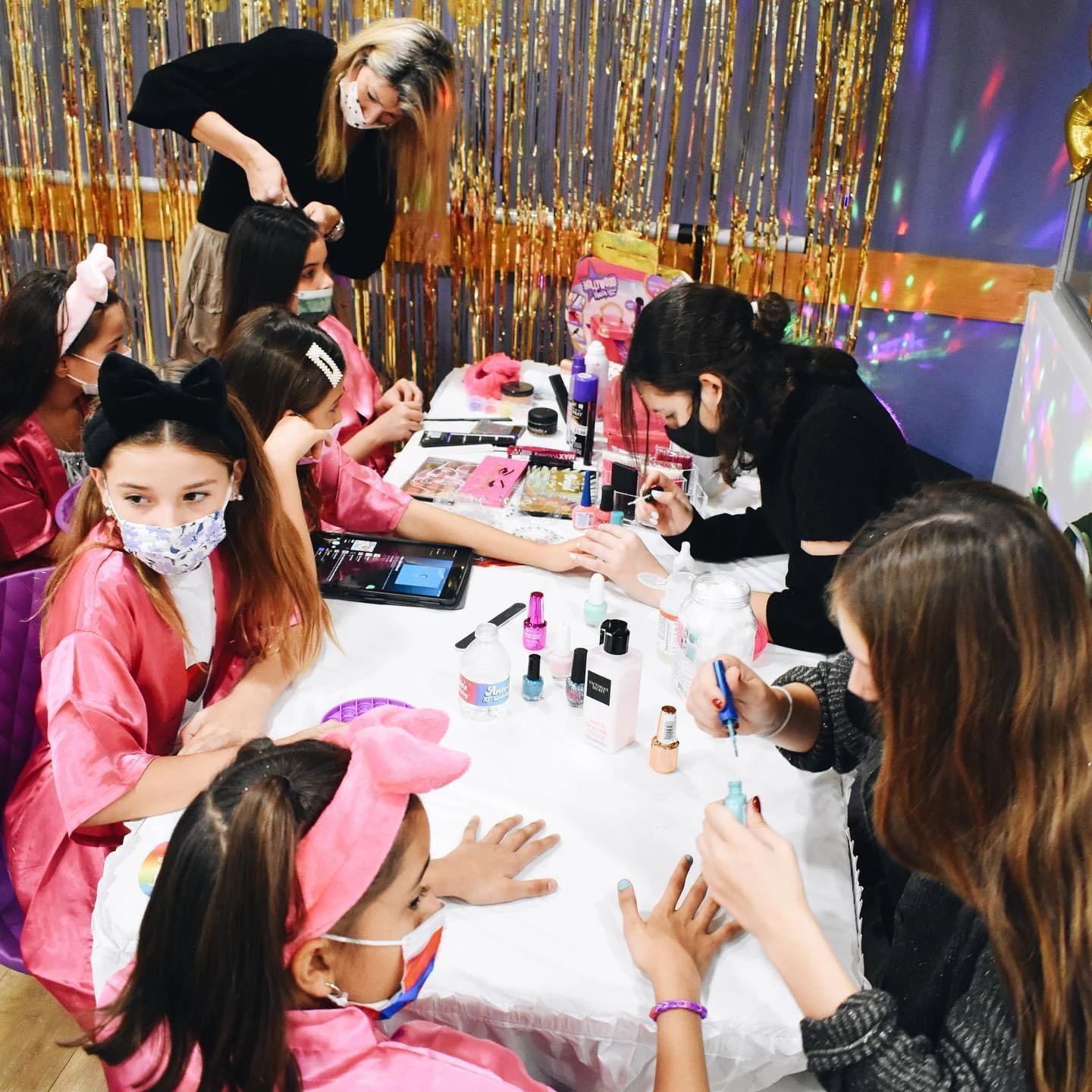 Group of girls in pink robes sit around a table having their nails painted while adults assist, surrounded by nail polish bottles and gold tinsel curtains
