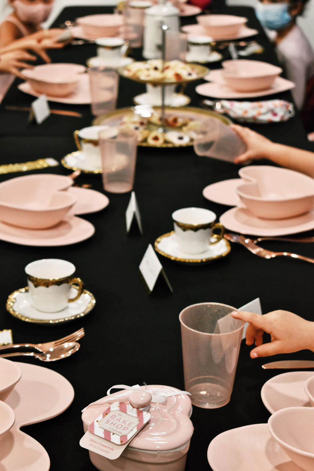 Children's hands reaching for glasses and place settings at a long black-draped table set with pink heart-shaped plates, gold-trimmed teacups, and a tiered tray of cookies