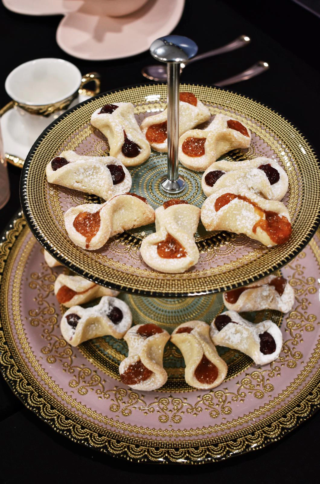 Two-tier gold and pink decorative stand holding powdered-sugar cookies filled with apricot and cherry jam