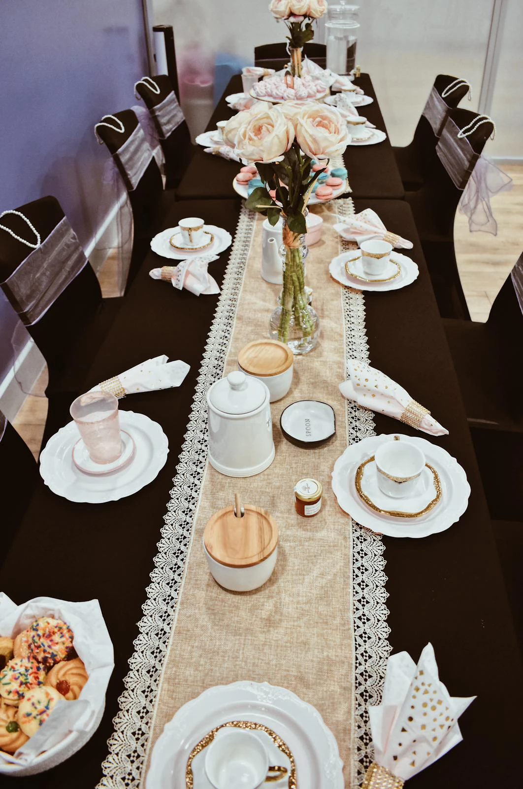 Elegant tea party table with a burlap and lace runner, white and gold teacups, folded gold-dotted napkins, pink roses, and a basket of sprinkle cookies