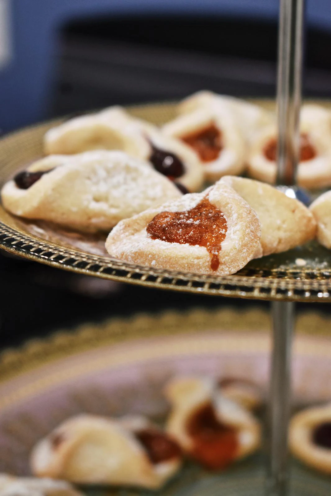 Close-up of a powdered-sugar cookie with apricot jam filling on a gold tiered stand, with more cookies blurred in the background