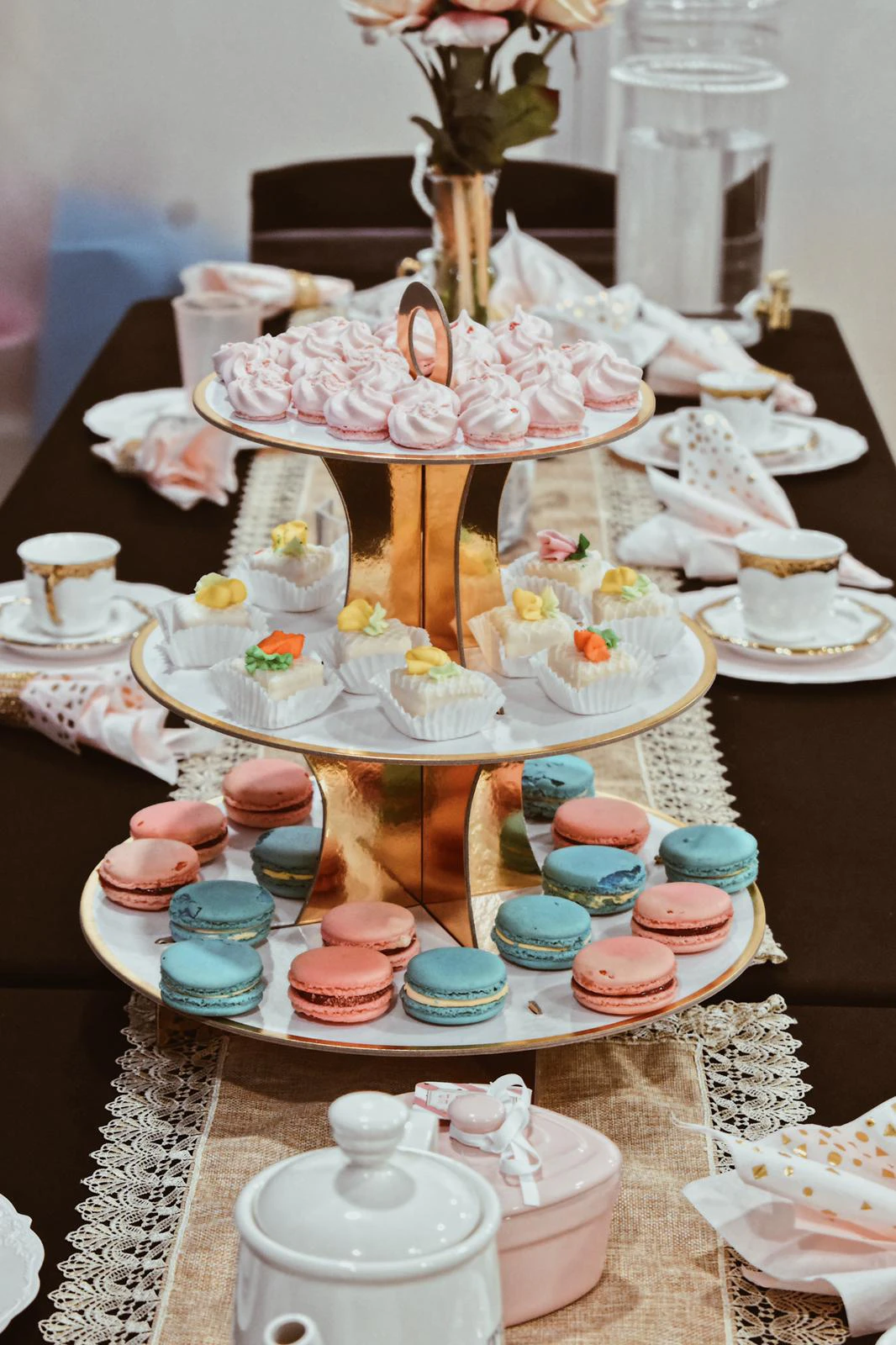 Three-tier gold and white dessert stand with pink meringues, small decorated petit fours, and pink and blue macarons on a tea party table