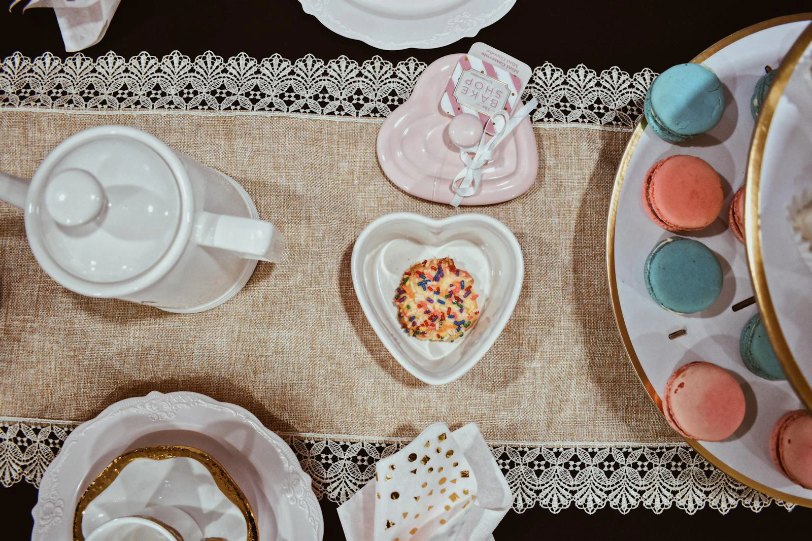 Overhead view of a tea table with a white teapot, a heart-shaped dish holding a sprinkle cookie, a pink heart-shaped casserole dish, and macarons on a lace-trimmed burlap runner