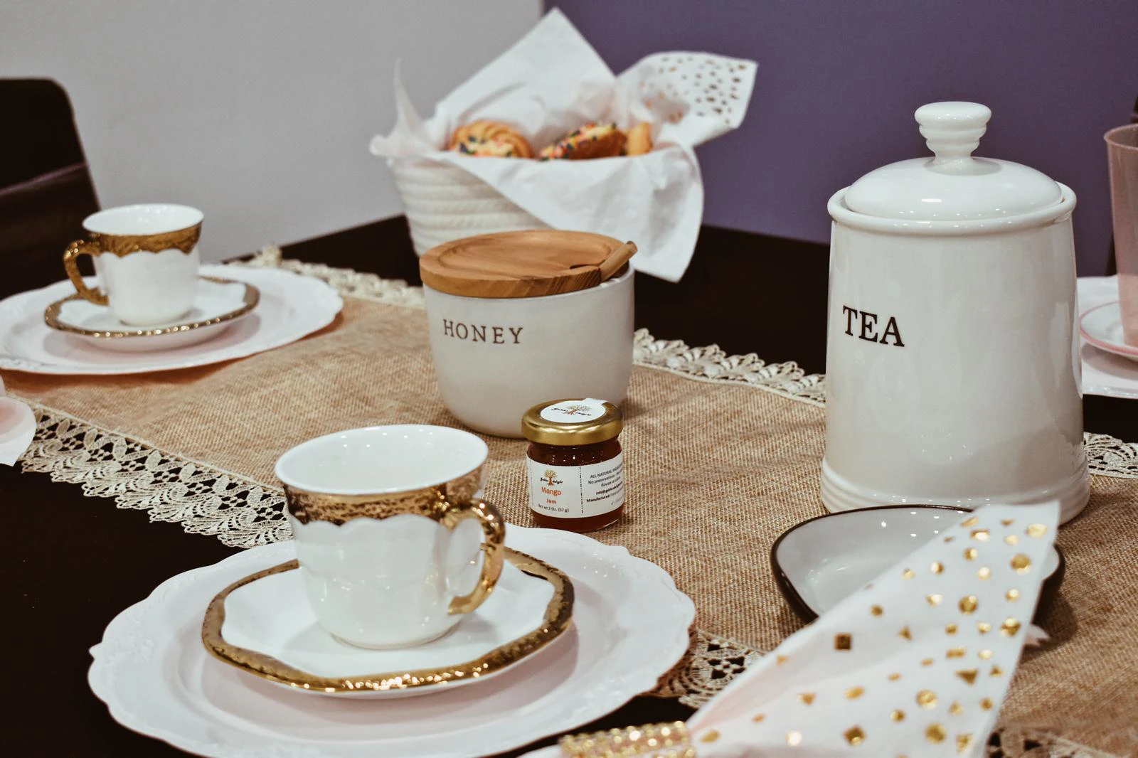 Gold-rimmed white teacup and saucer on a plate beside jars labeled Tea and Honey, a small jar of mango jam, and a basket of cookies