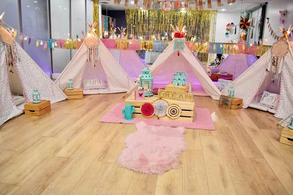 Colorful indoor teepee sleepover setup with patterned tents, pink faux-fur rug, wooden crates spelling “DREAM,” lanterns, dreamcatchers, and a “Wild Child” banner