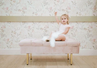 Adorable toddler girl in a pale pink leotard and ballet slippers sitting on a blush-pink velvet bench with gold legs, wearing a white feather boa and raising one arm, against floral wallpaper.