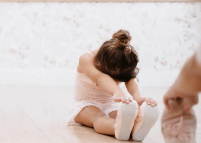 Young girl in a light pink leotard sitting on the wooden dance floor, bending forward with hands reaching toward her pointed ballet slippers, hair in a neat bun.