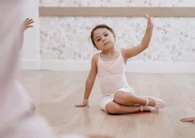 Young girl in a pale pink camisole leotard and ballet slippers sitting cross-legged on the studio floor, one arm gracefully raised, looking toward the camera with floral wallpaper in the background.